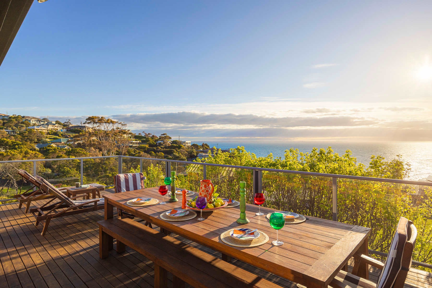 A timber table set for dinner on a deck above the bay, the sun setting over the water