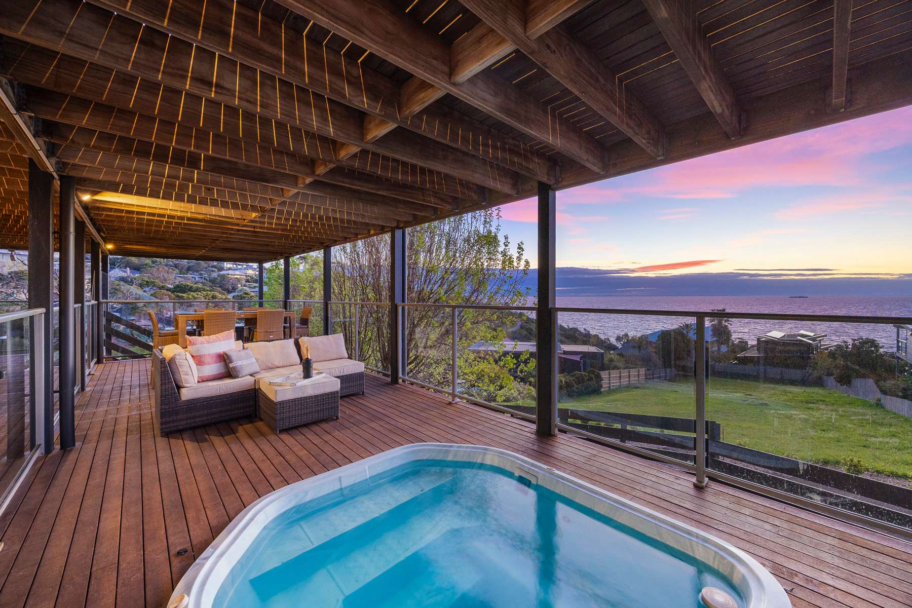 A spa set into a covered timber deck with outdoor sofas, looking out to the bay under a pink sunset sky