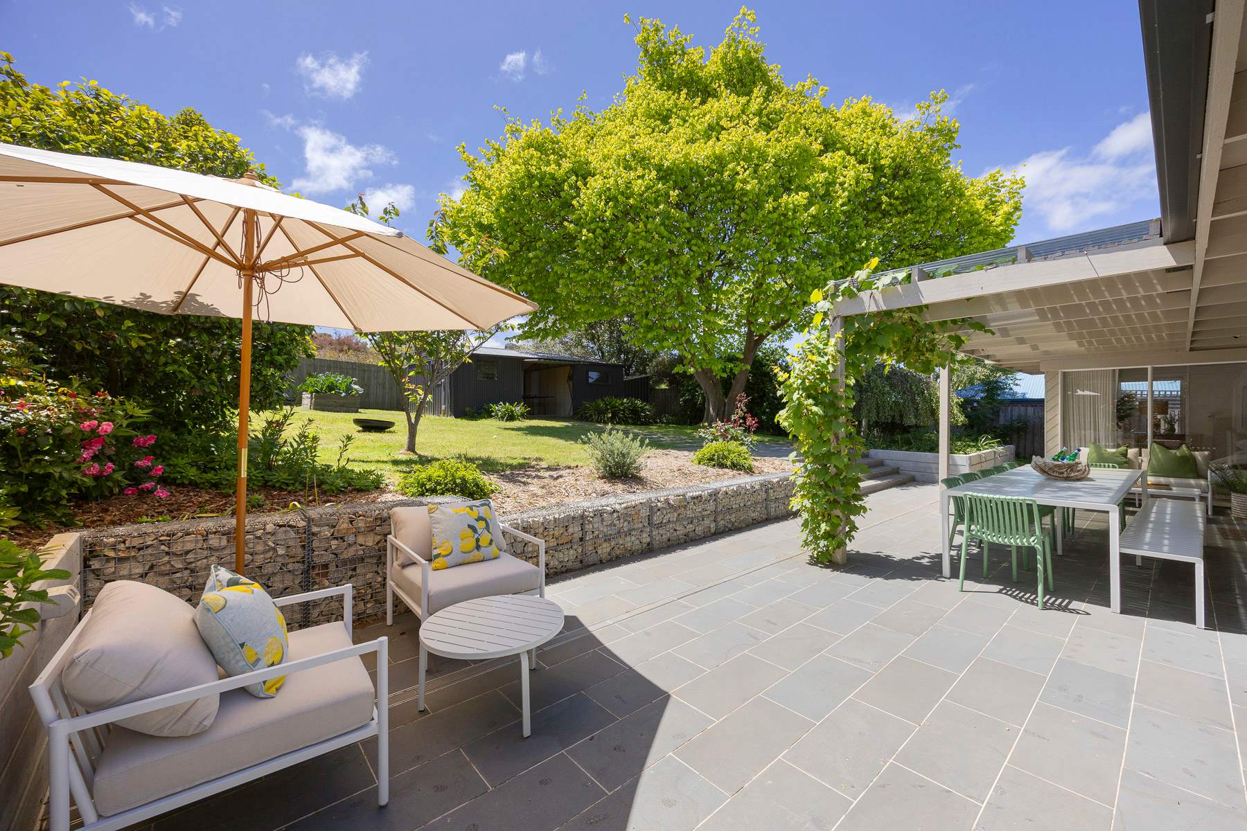 A sunny garden terrace with an umbrella, outdoor seating and a long table among green trees