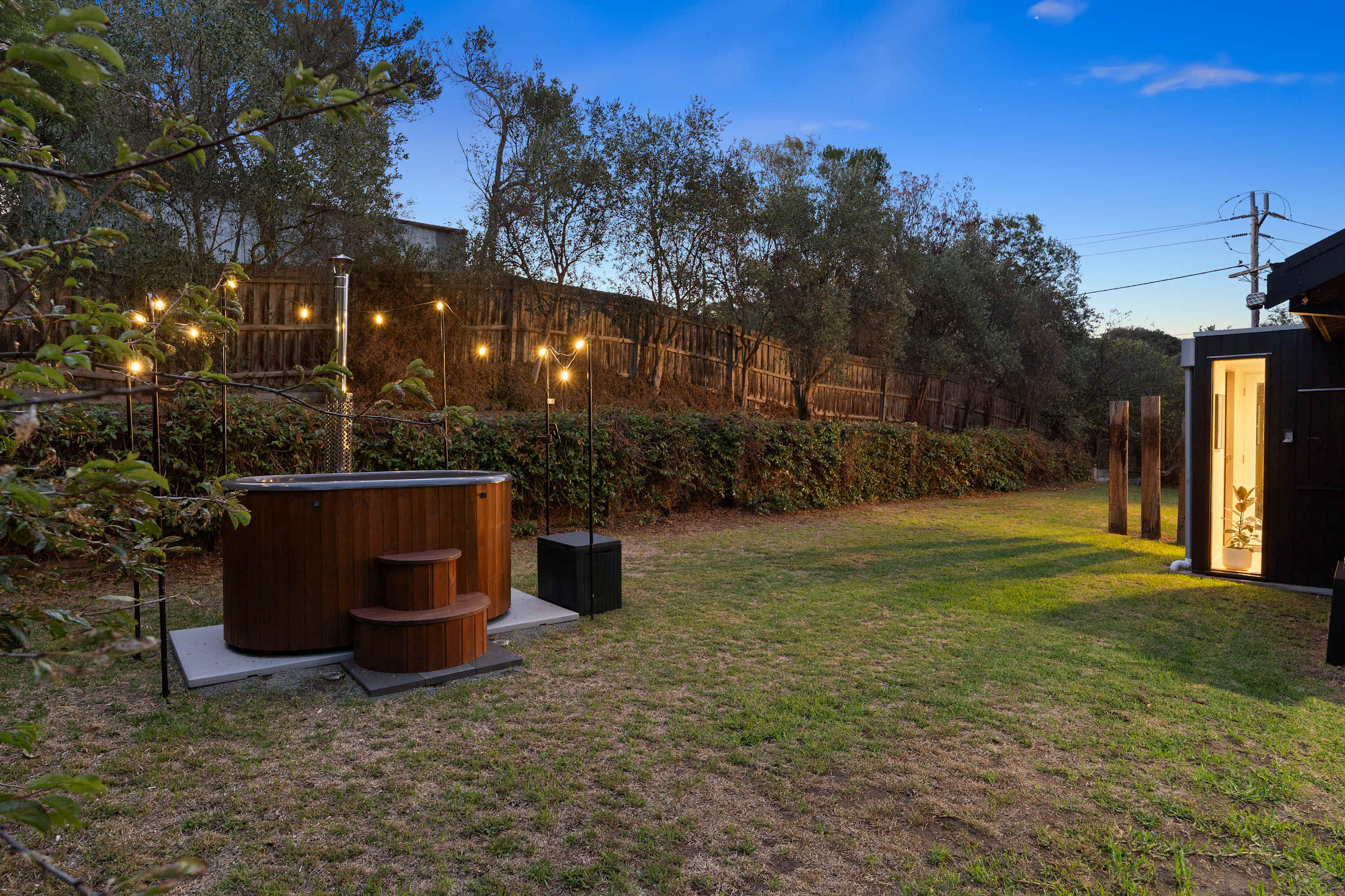 A round timber hot tub on a lawn at dusk under festoon lights, beside a small black studio with a lit doorway