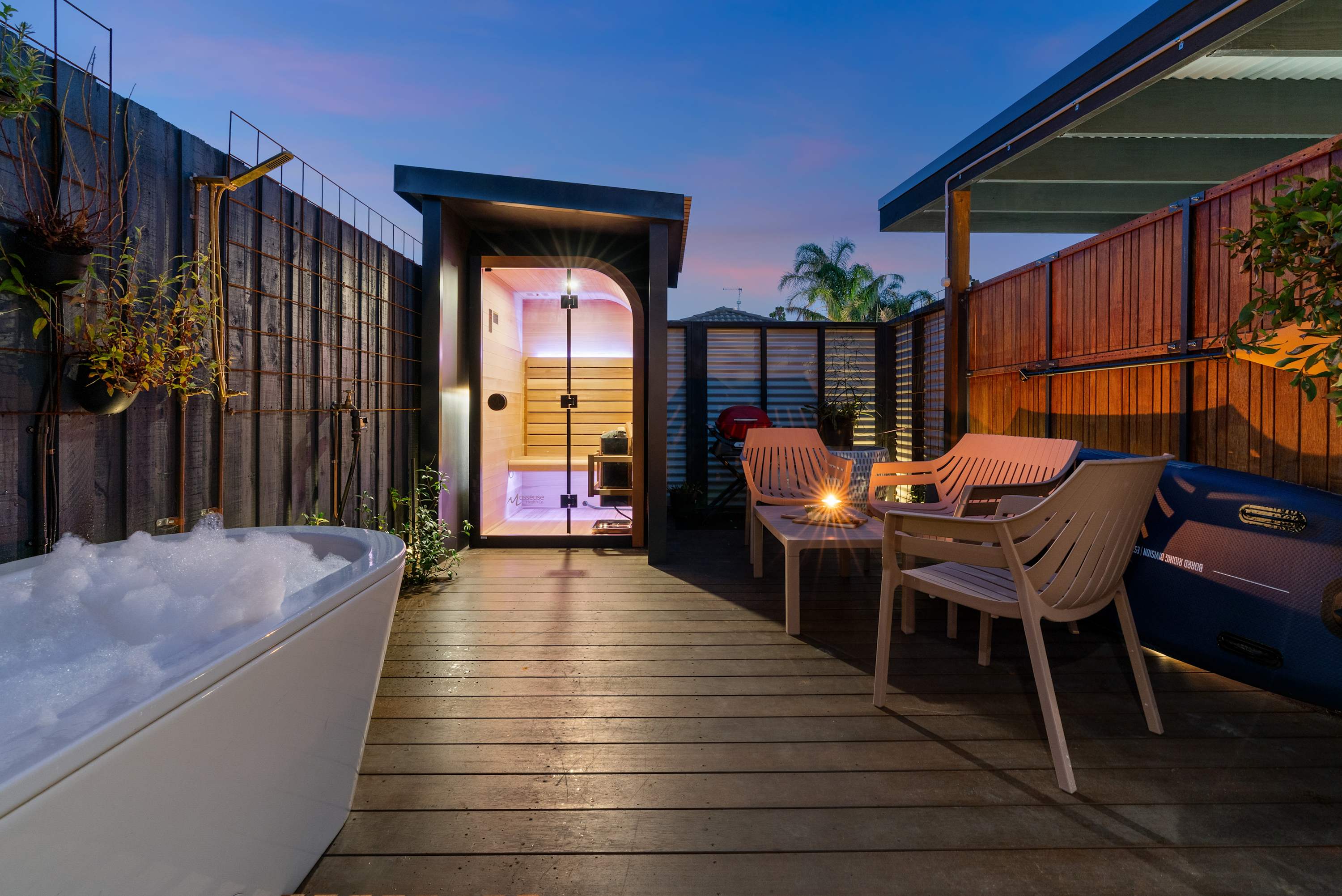 A freestanding bath full of bubbles on a timber deck at dusk, with a glowing glass-fronted sauna behind