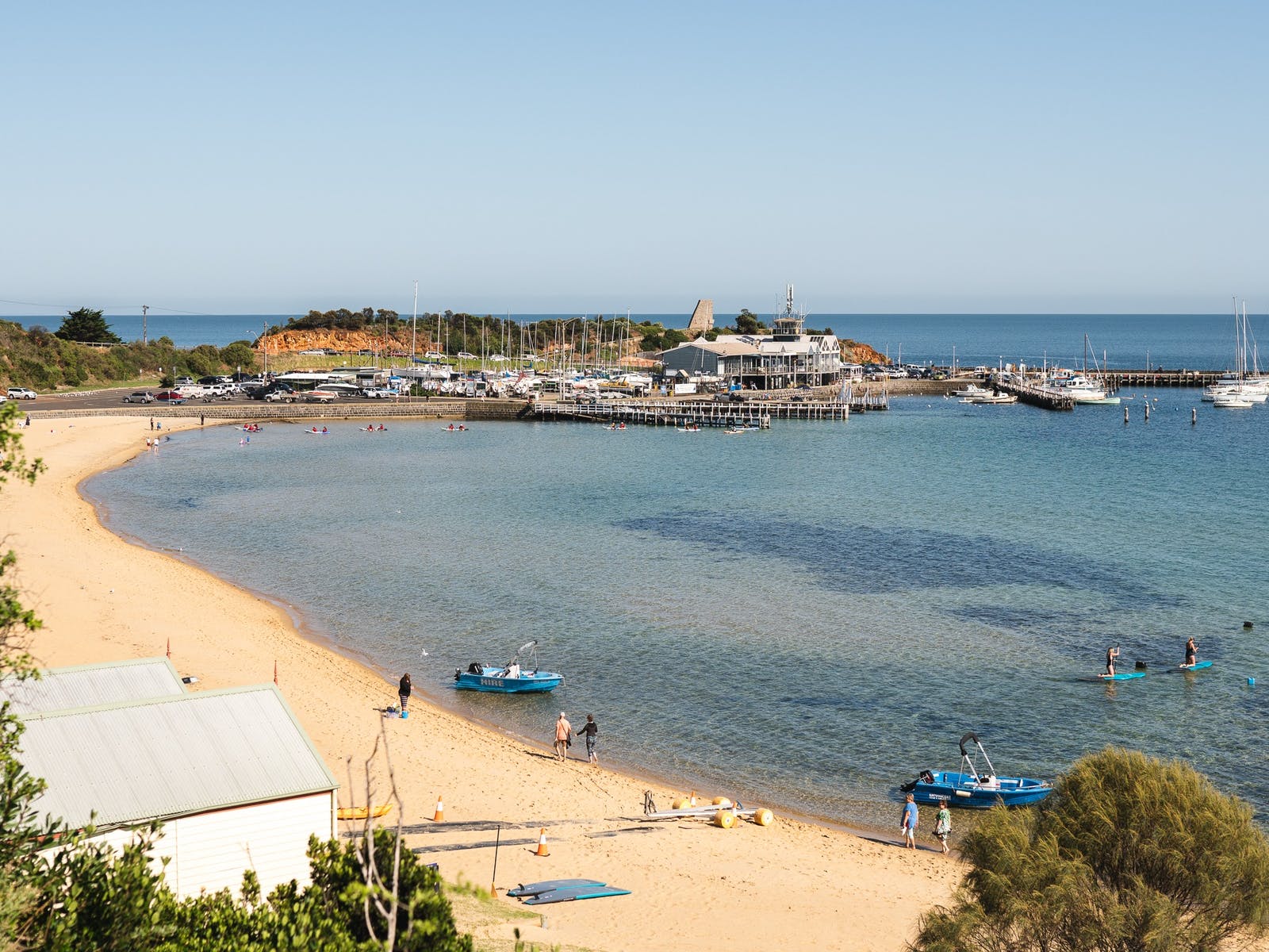 A curving sandy beach on calm water, with boats moored off a pier and a harbour beyond