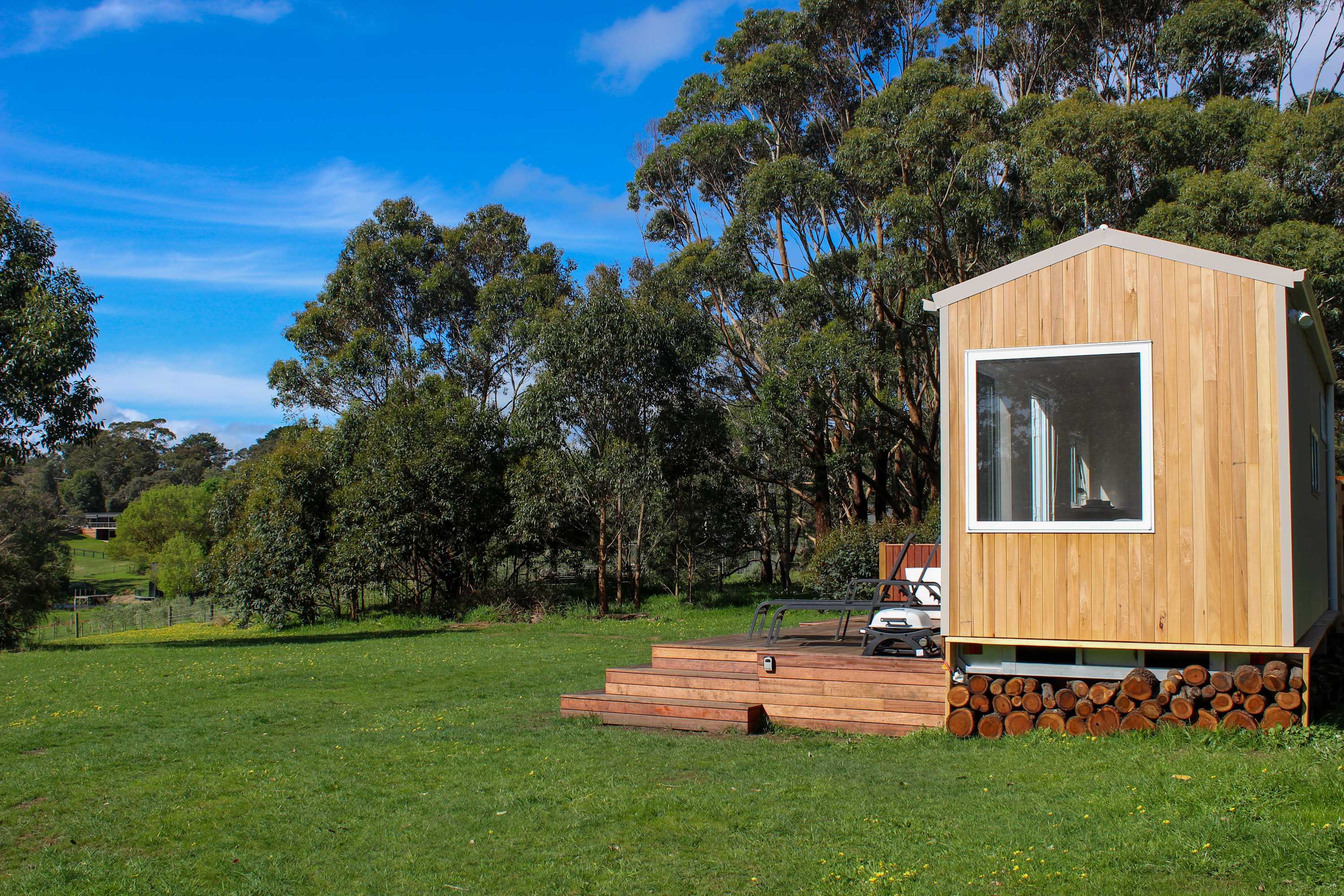 A small timber cabin with a deck and stacked firewood in a green paddock fringed by tall gum trees